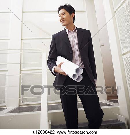 Stock Image - Low angle view of a businessman holding blueprints and smiling. Fotosearch