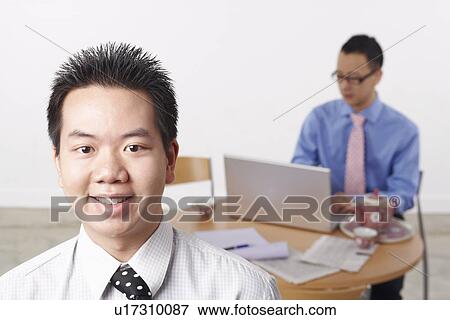 Stock Photo - Portrait of a businessman smiling with another businessman using a laptop behind him. Fotosearch
