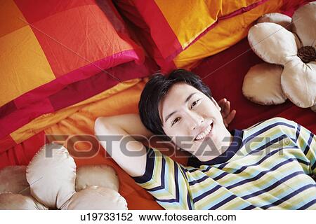 Stock Image - Portrait of a young man smiling and lying on the bed with his hand behind his head. Fotosearch
