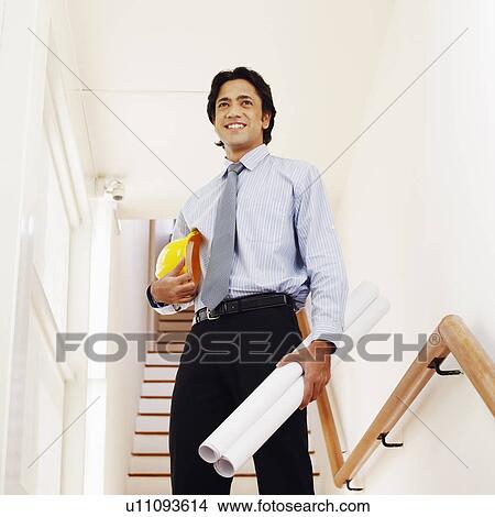 Picture - Low angle view of a male architect holding rolled up blueprints and a hardhat. Fotosearch