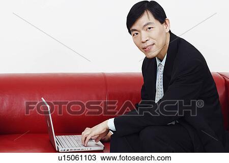Stock Image - Portrait of a businessman sitting on a couch and using a laptop. Fotosearch