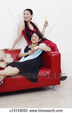 Stock Image - Two mid adult women sitting on a couch with champagne flutes and smiling. Fotosearch