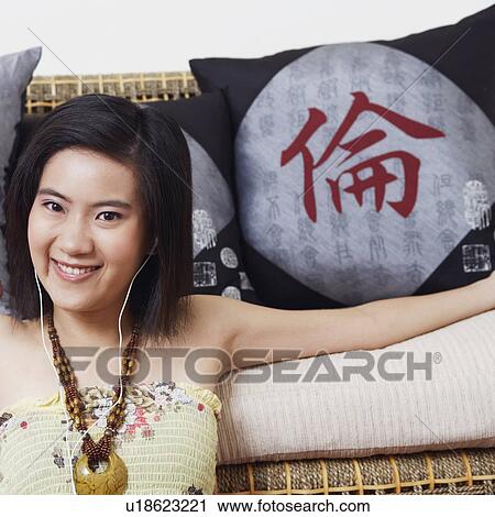 Stock Image - Portrait of a young woman leaning against a couch and listening to music. Fotosearch