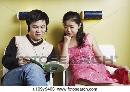 Stock Image - Young man and a mid adult woman sitting on chairs listening to music. Fotosearch