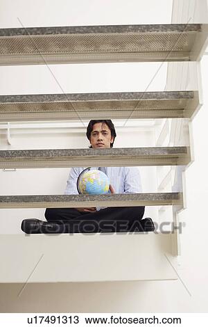 Stock Image - Portrait of a businessman sitting cross-legged on a step and holding a globe. Fotosearch
