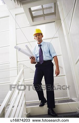 Stock Image - Portrait of an architect walking down stairs and carrying blueprints. Fotosearch