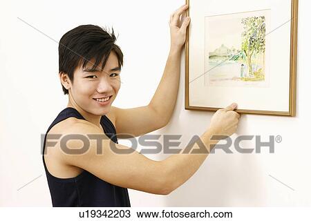 Stock Image - Portrait of a young man hanging a picture frame on wall and smiling. Fotosearch