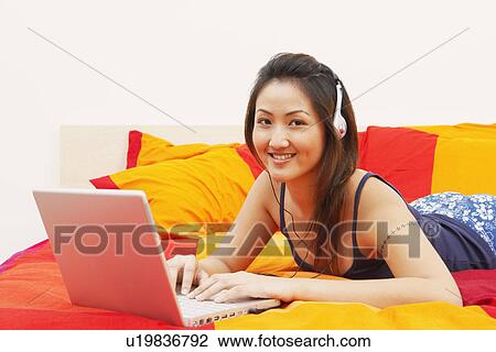 Stock Image - Portrait of a young woman lying on the bed and using a laptop. Fotosearch