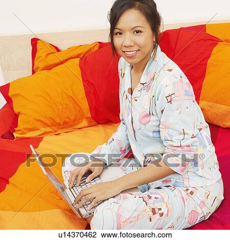 Stock Image - Portrait of a young woman sitting on the bed and using a laptop. Fotosearch