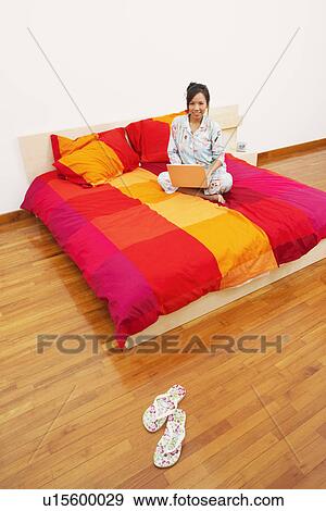 Portrait of a young woman sitting on the bed and using a laptop View Large Photo Image Stock Photo - Portrait of a young woman sitting on the bed and using a laptop. Fotosearch