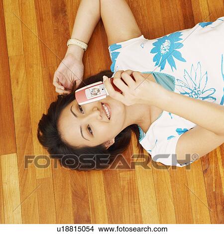 Picture - High angle view of a young woman lying on a hardwood floor and holding a mobile phone. Fotosearch