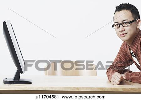 Stock Image - Portrait of a businessman leaning against a table in front of a flat screen monitor. Fotosearch