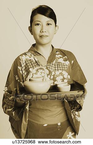 Stock Photo - Portrait of a young woman holding tea cups and a tea kettle on a tray. Fotosearch