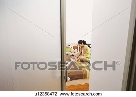 Stock Image - High angle view of a young woman sitting on the bed and using a laptop. Fotosearch
