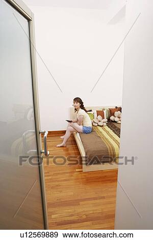 Stock Photo - Portrait of a young woman sitting on the bed and holding a mobile phone. Fotosearch