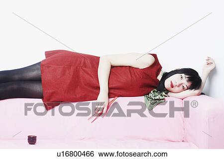 Stock Photograph - Portrait of a young woman lying on a couch holding chopsticks. Fotosearch