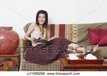 Stock Photograph - Portrait of a young woman sitting on a couch and holding a magazine. Fotosearch