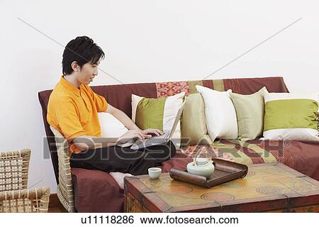 Stock Photograph - Side profile of a young man sitting on a couch and using a laptop. Fotosearch
