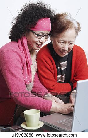 Stock Photo - Side profile of a mature woman sitting with her friend using a laptop. Fotosearch