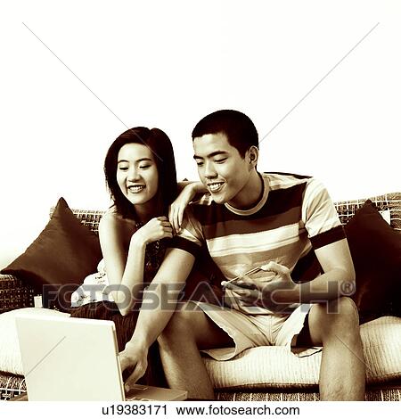 Stock Image - Young woman sitting beside a young man holding a credit card and using a laptop. Fotosearch