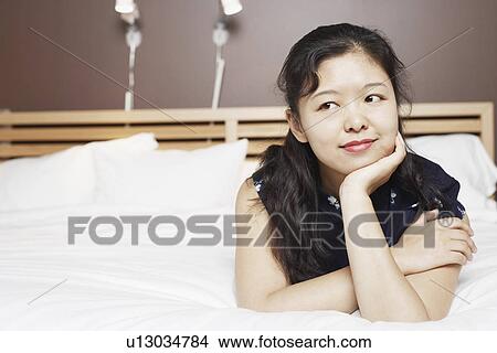 Picture - Close-up of a young woman lying on the bed thinking with her hand on her chin. Fotosearch