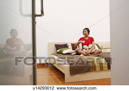 Young woman sitting on the bed and holding a cup of tea View Large Photo Image Stock Image - Young woman sitting on the bed and holding a cup of tea. Fotosearch