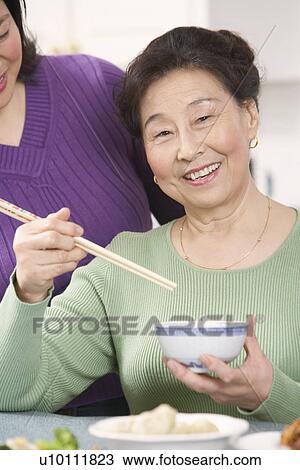 Portrait of a senior woman eating food with her daughter standing behind her View Large Photo Image Stock Image - Portrait of a senior woman eating food with her daughter standing behind her. Fotosearch