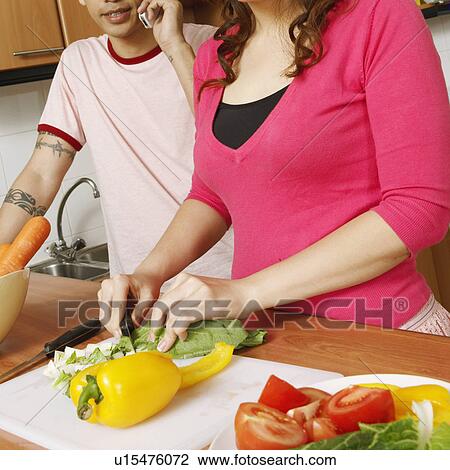 Stock Image - Portrait of a young woman cutting vegetables with a mid adult man talking on a mobile phone beside her. Fotosearch