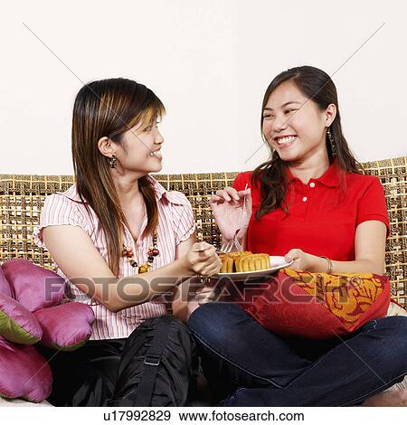 Stock Photo - Two young women sitting together on a couch and looking cheerful. Fotosearch