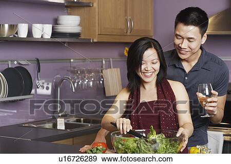Stock Photo - Young man standing holding a wineglass with a young woman preparing food. Fotosearch