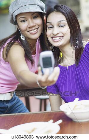 Stock Image - Close-up of two young women taking a photograph of themselves with a mobile phone. Fotosearch