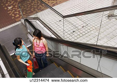 Picture - High angle view of two young women standing on an escalator with shopping bags. Fotosearch