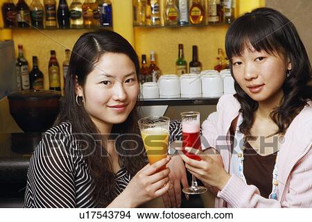 Picture - Portrait of two young women sitting at a bar counter raising a toast. Fotosearch