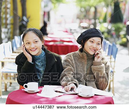 Stock Photo - Two young woman sitting in a restaurant and talking on mobile phones. Fotosearch
