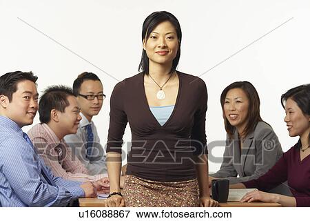 Stock Photo - Portrait of a businesswoman leaning against a table with business executives sitting behind her. Fotosearch