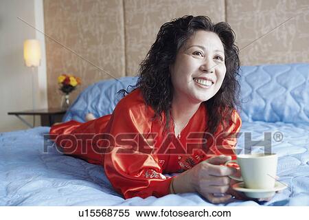 Stock Photography - Mature woman lying on the bed with a cup of tea smiling. Fotosearch