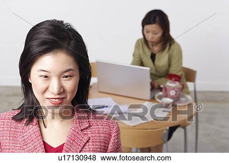 Stock Photo - Portrait of a businesswoman smiling with another businesswoman using a laptop behind her. Fotosearch