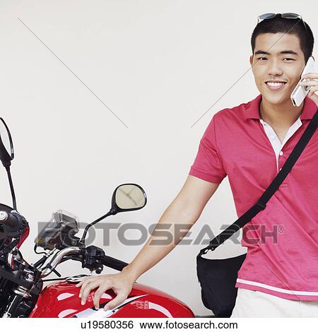 Stock Photograph - Close-up of a young man talking on a mobile phone and leaning against a motorcycle. Fotosearch