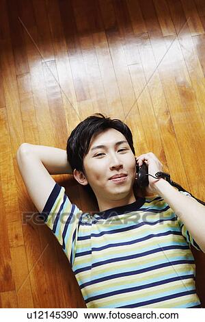 Stock Image - High angle view of a young man talking on the telephone and lying on a hardwood floor. Fotosearch