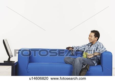 Stock Image - Side profile of a young man holding a remote control sitting in front of a flat screen monitor. Fotosearch