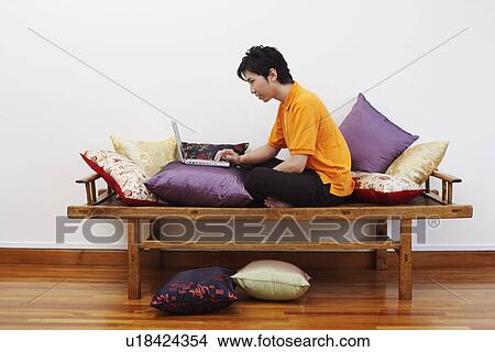 Picture - Side profile of a young man sitting on a bench and using a laptop. Fotosearch