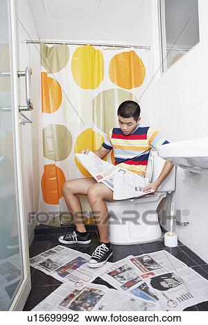 Stock Image - Young man sitting on a toilet bowl and reading a newspaper. Fotosearch