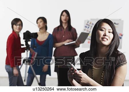 Portrait of a young woman holding a mobile phone with her friends standing behind her View Large Photo Image Picture - Portrait of a young woman holding a mobile phone with her friends standing behind her. Fotosearch