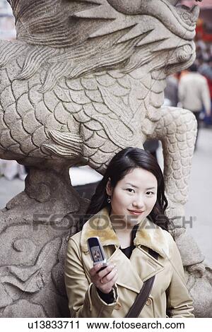 Stock Image - Portrait of a young woman leaning against a statue holding a mobile phone. Fotosearch