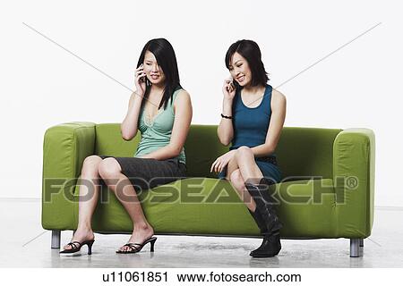 Two young women sitting on a couch talking on mobile phones View Large Photo Image Stock Image - Two young women sitting on a couch talking on mobile phones. Fotosearch
