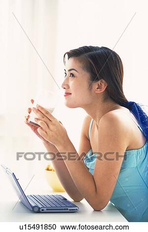 Stock Image - Image of a Mid Adult Woman Drinking a Cup While Sitting in Front of her Laptop, Smiling, Looking Sideways, Side View. Fotosearch
