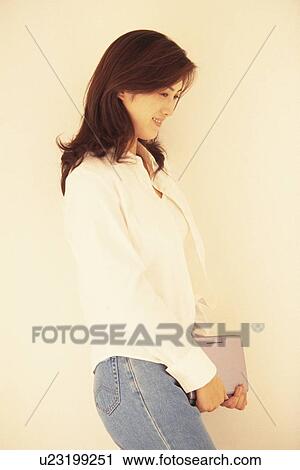 Stock Image - Image of a Young Adult Woman Holding a Laptop, Standing In Front of a White Wall, Side View. Fotosearch