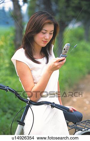 Picture - Image of a Young Adult Woman Looking at her Cell Phone, While Holding her Bicycle, Side View. Fotosearch