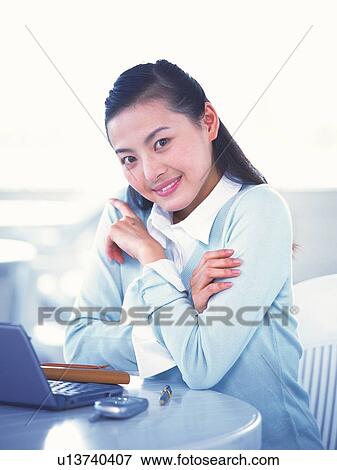 Stock Photo - Image of a Young Adult Woman Sitting at a Table In Front of a Laptop, Smiling, Side View, Looking at Camera. Fotosearch