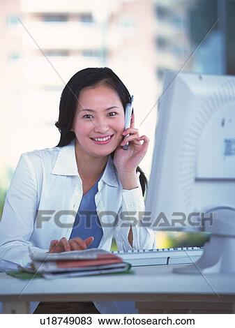 Stock Image - Image of a Young Businesswoman Working at her Desk While Talking on a Cell phone, Smiling, Front View. Fotosearch